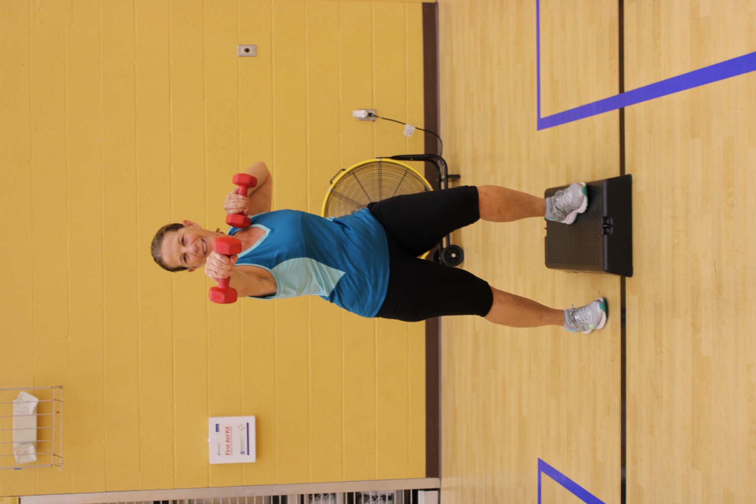 A happy woman with pink weights in her hands, looking at the camera with one foot up on a black stepper